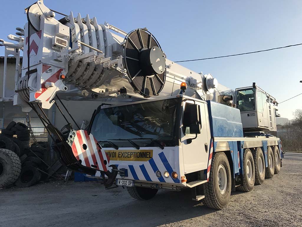 Gros camion-grue blanc robuste stationné à l'extérieur sur un terrain en terre battue, avec une flèche levée et un pare-chocs avant rayé.