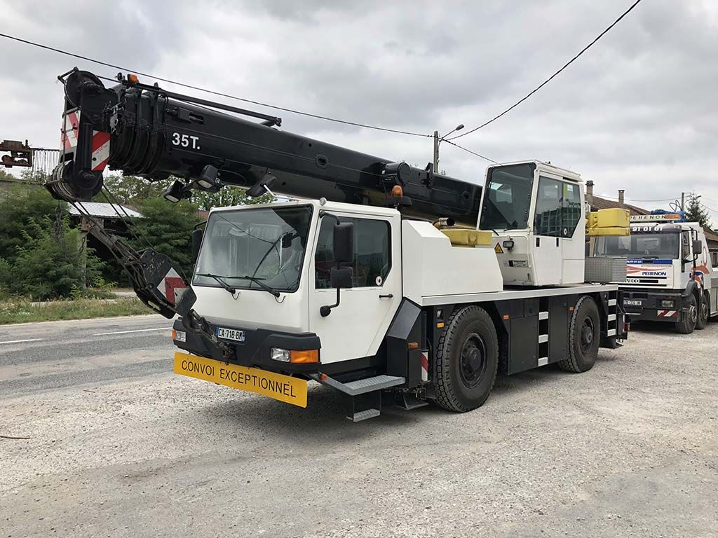 Camion-grue mobile blanc stationné sur un terrain gravillonné, flèche noire déployée sous un ciel nuageux