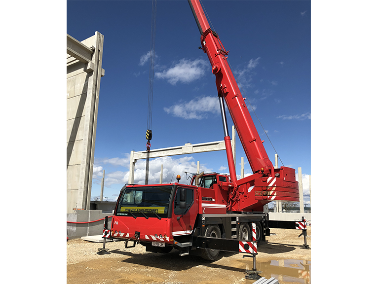 Une grue mobile rouge soulève une charge à côté d'un camion rouge sous un ciel bleu sur un chantier de construction