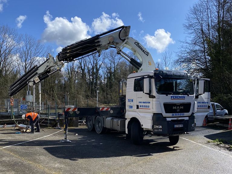 Camion-grue blanc déchargeant des matériaux sur un chantier en bord de route, un ouvrier à proximité.