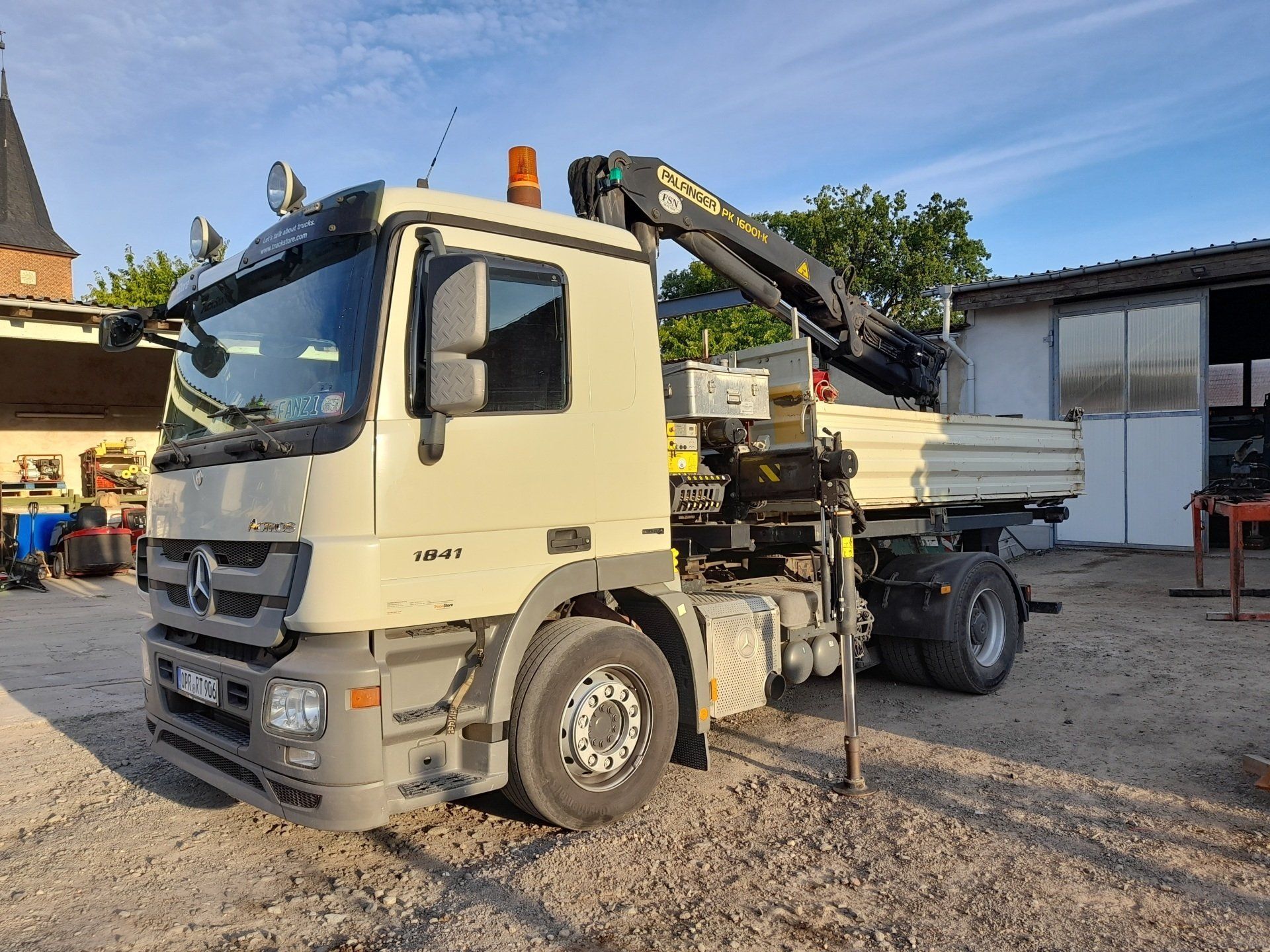 Ein weißer Lastwagen mit einem Kran auf der Ladefläche steht auf einem Schotterplatz.