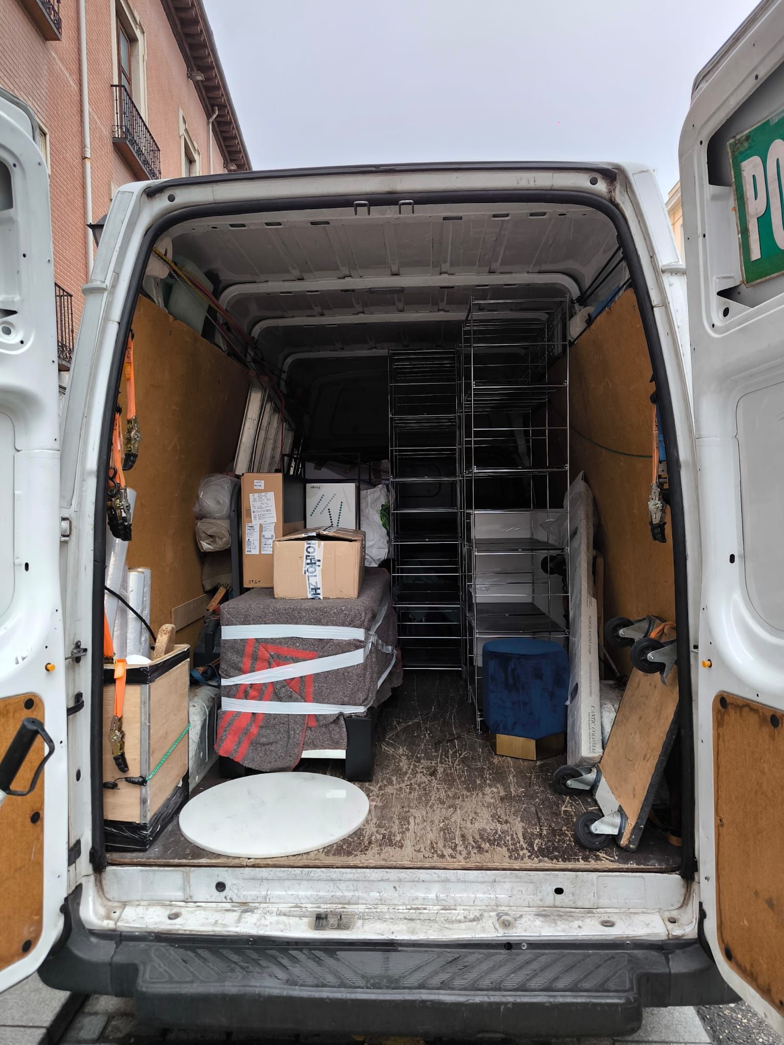 Parte trasera abierta de una furgoneta blanca llena de diversos artículos, incluyendo cajas y estanterías. La vista interior muestra la furgoneta estacionada en la calle.