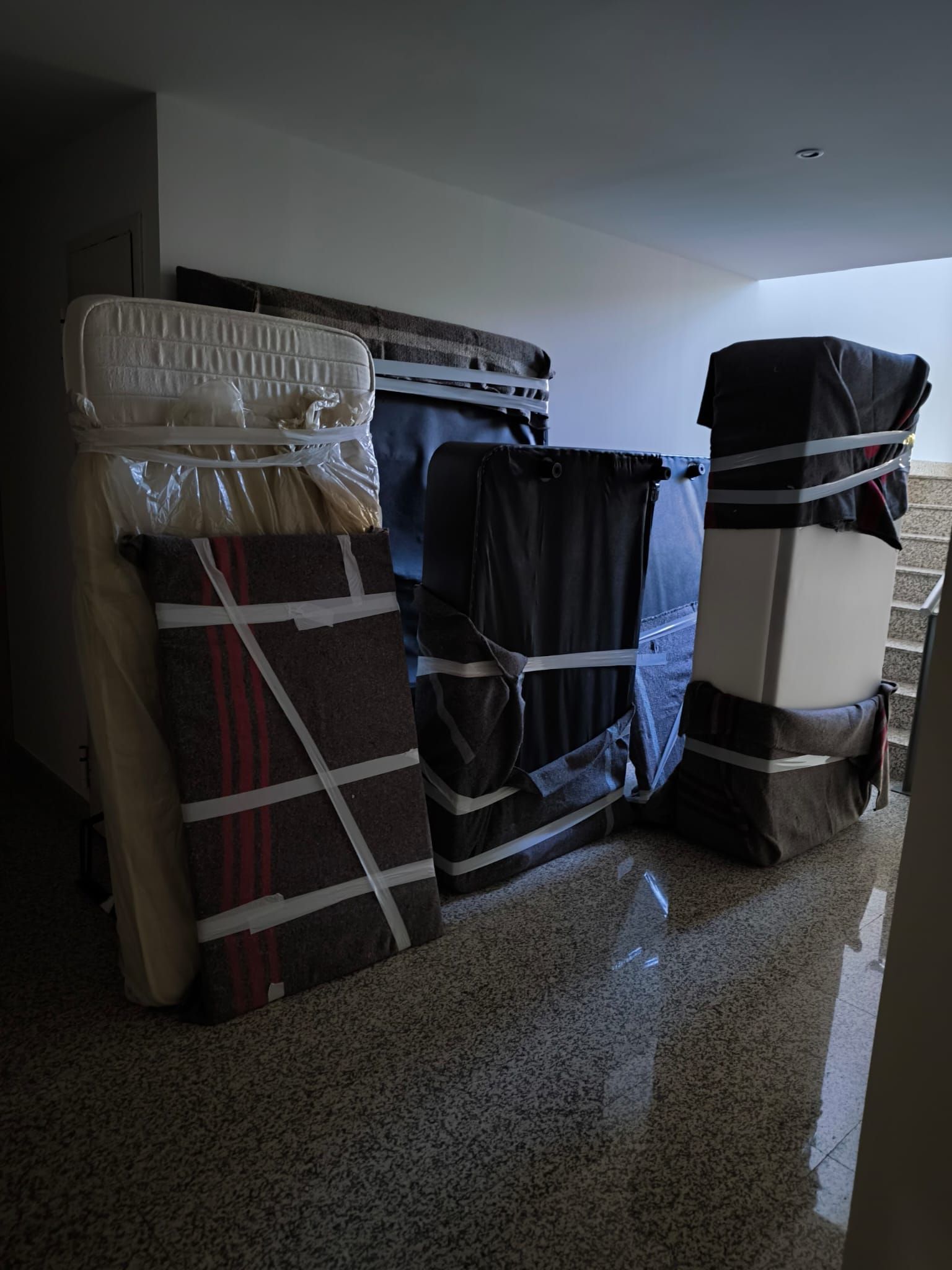 Muebles envueltos y apilados en un pasillo. Colchones y cajas cubiertos con plástico y asegurados con cinta adhesiva, cerca de las escaleras.