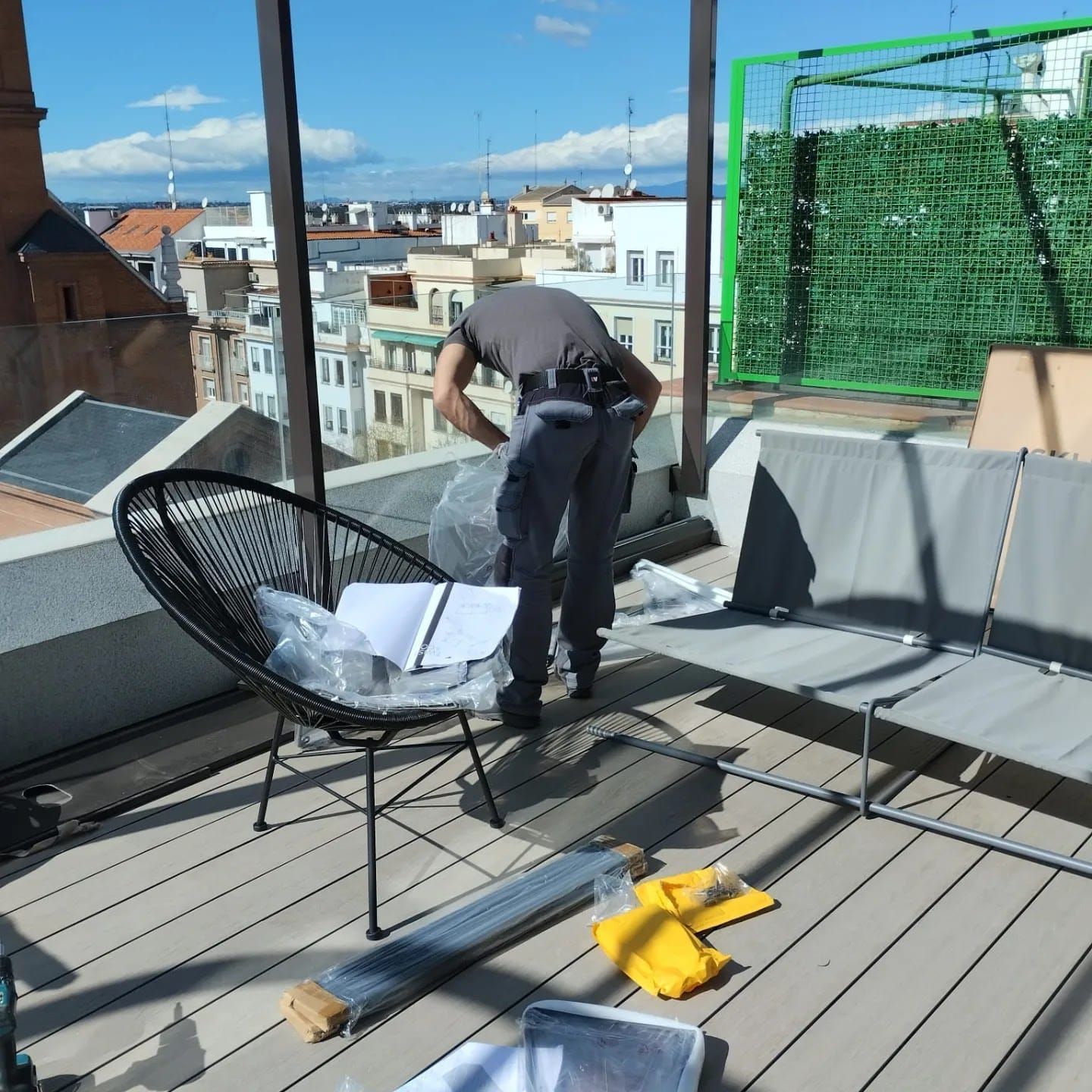 Una persona desempaca muebles en una terraza con vistas a la ciudad. También hay muebles grises y negros y una pantalla verde.