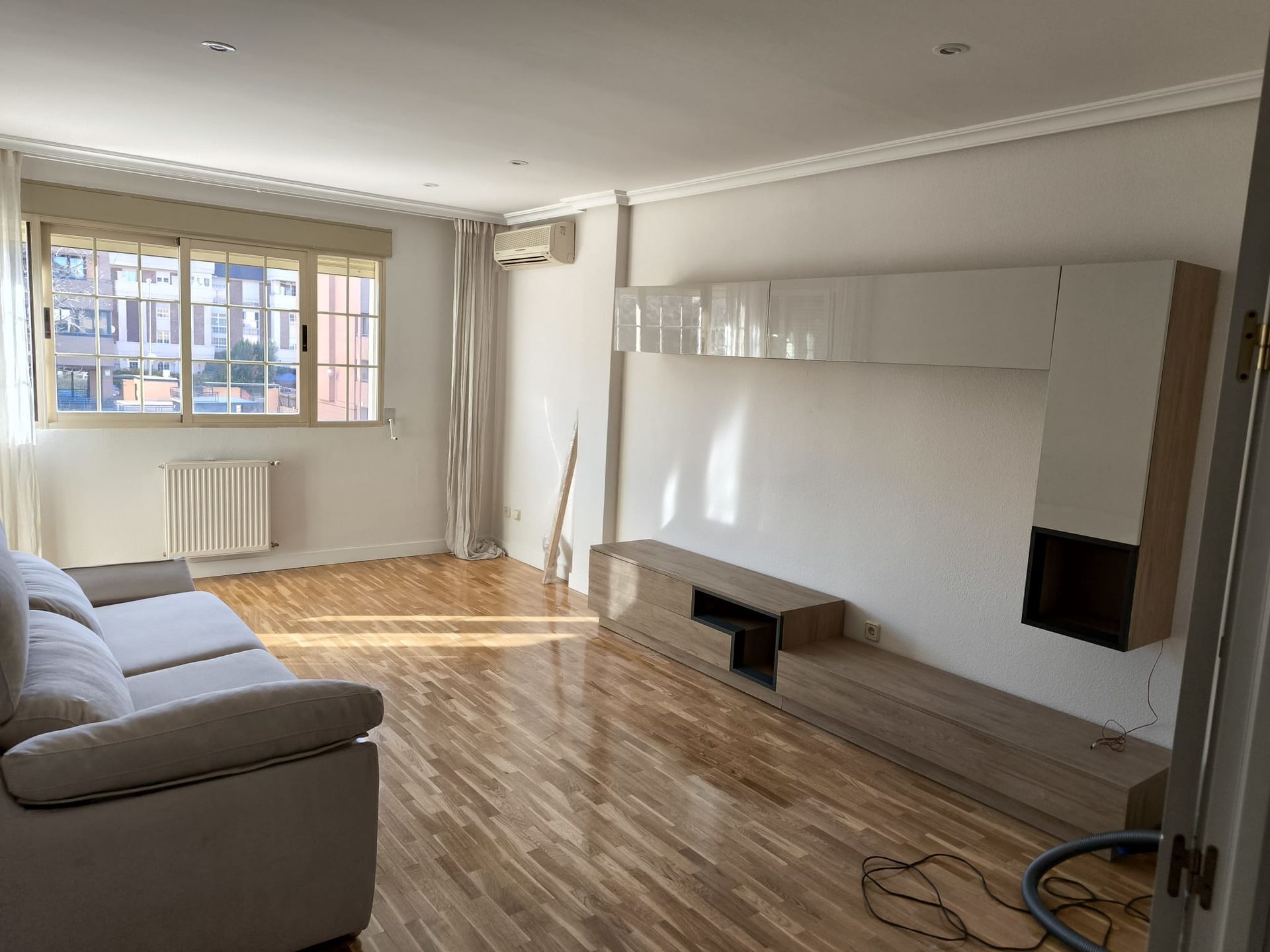 Una sala de estar moderna y vacía con un sofá blanco, suelo de madera y un sistema de entretenimiento integrado. La luz natural entra a raudales por una ventana.