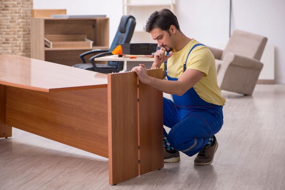 Un hombre con overol azul ensambla un escritorio de madera en una habitación. Se arrodilla, usando una herramienta en un panel lateral del escritorio.