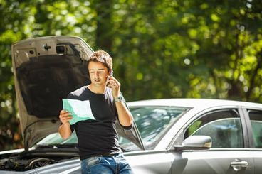 Un hombre de pie junto a un coche plateado con el capó abierto, sujetando unos papeles y usando el teléfono al aire libre, bajo la luz del sol