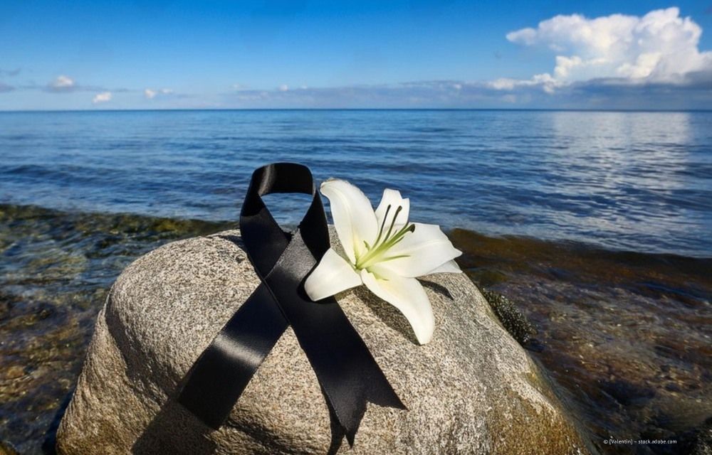 Schwarzes Trauerband und weiße Lilie auf einem Felsen am Meer unter blauem Himmel.