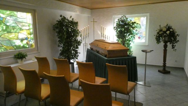 Eine brennende weiße Kerze neben einem weißen Blumenarrangement auf einem Holzsarg in einem schwach beleuchteten Raum