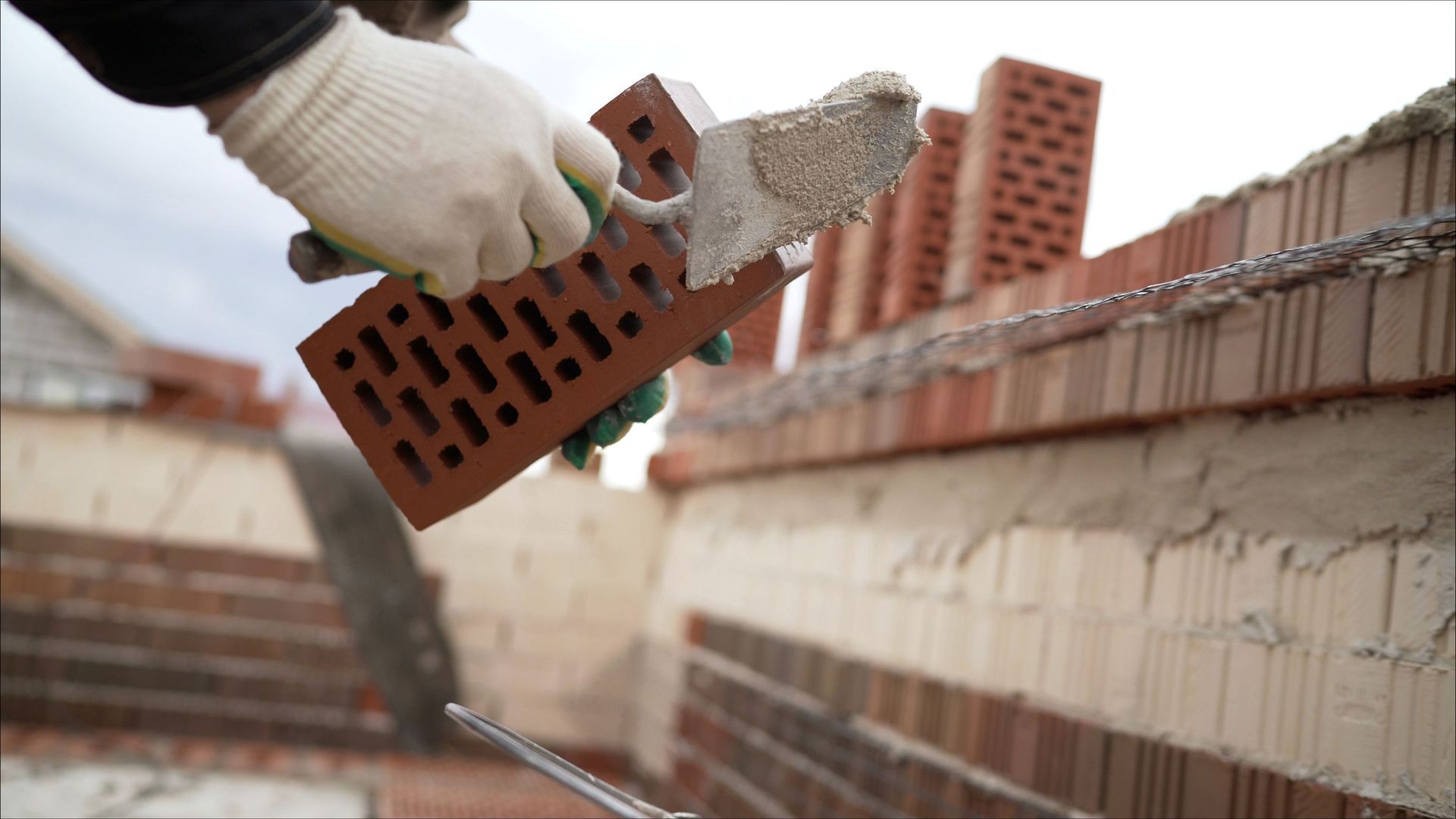 Persona con guantes colocando un ladrillo rojo, utilizando una paleta y mortero, en un sitio de construcción.