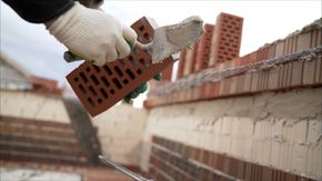 Persona con guantes colocando un ladrillo rojo, utilizando una paleta y mortero, en un sitio de construcción.