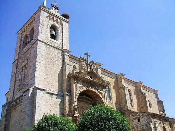 Iglesia de piedra con campanario, cruz y cigüeñas en un día soleado.