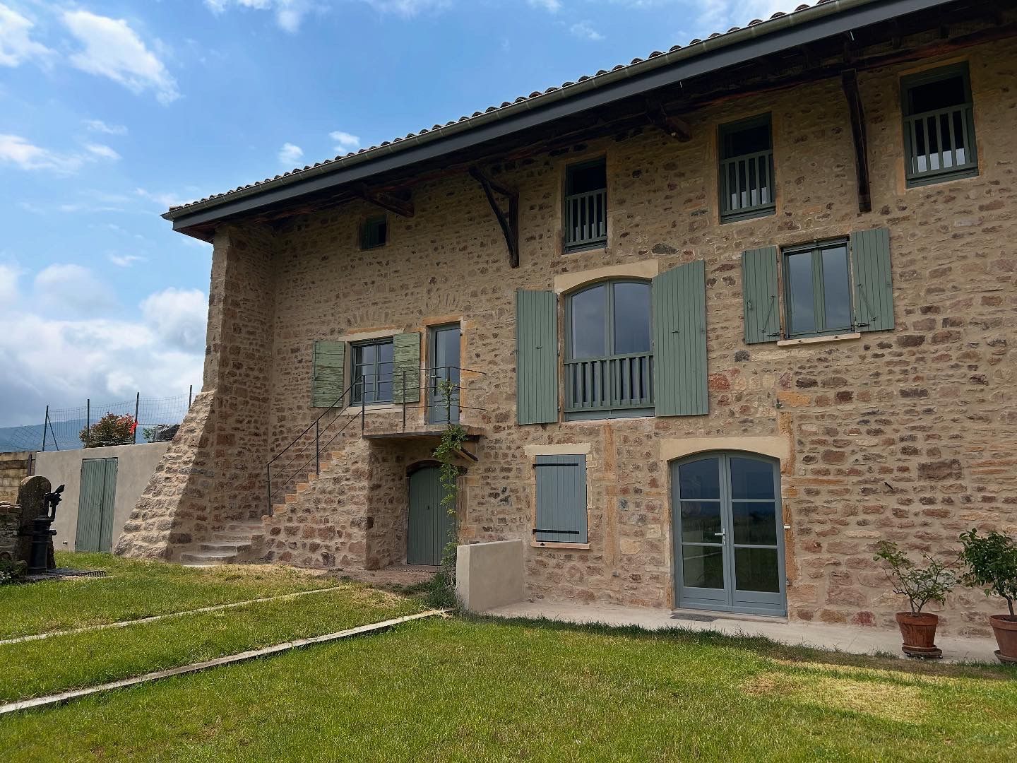 Maison en pierre avec volets et portes verts, située dans une cour herbeuse sous un ciel bleu.