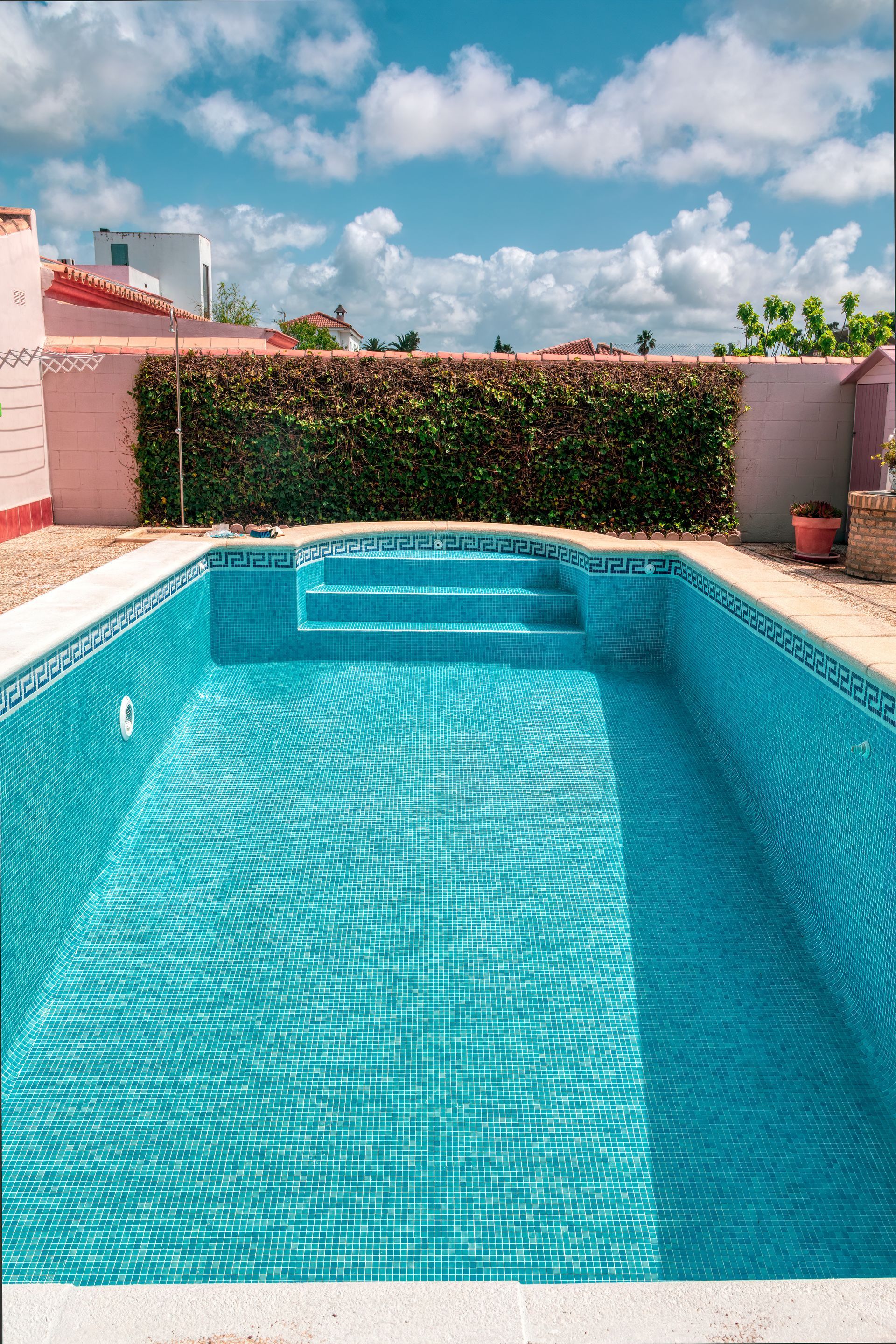 Une piscine carrelée turquoise avec marches, face à une haie verte, sous un ciel bleu nuageux.