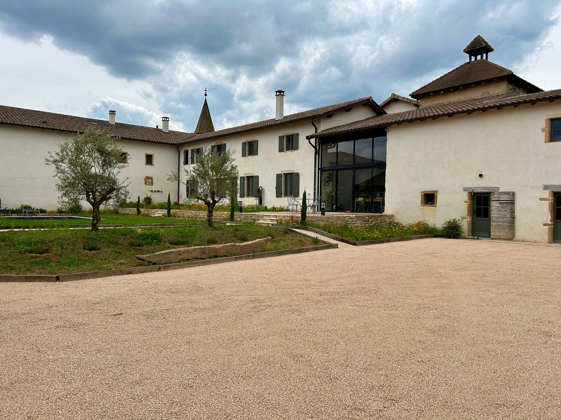 Un grand bâtiment blanc avec une cour en gravier et un petit jardin. Ciel couvert.