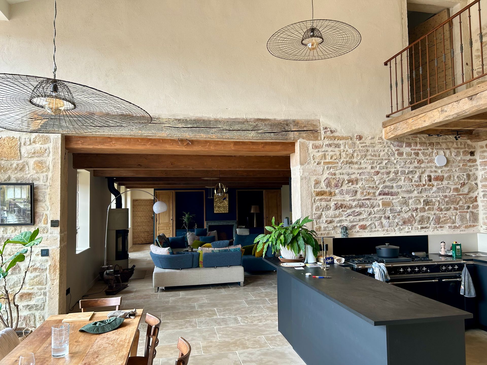 Séjour ouvert avec murs en pierre, îlot central et mezzanine. Meublé avec canapés bleus, plantes et poutres apparentes.