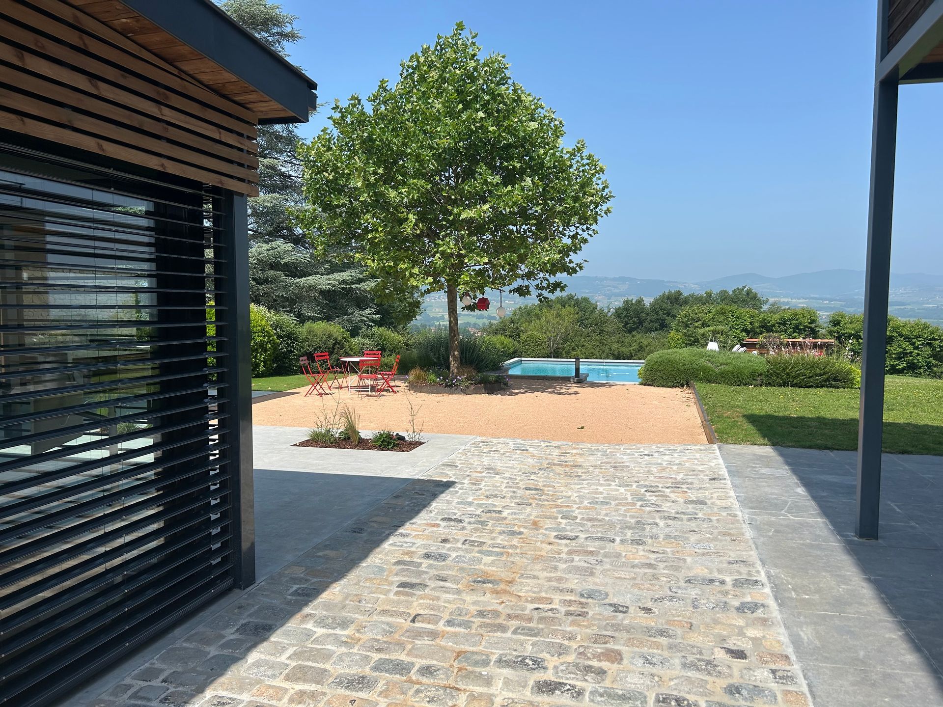 Vue depuis un patio vers une piscine et un paysage sous un ciel bleu clair.