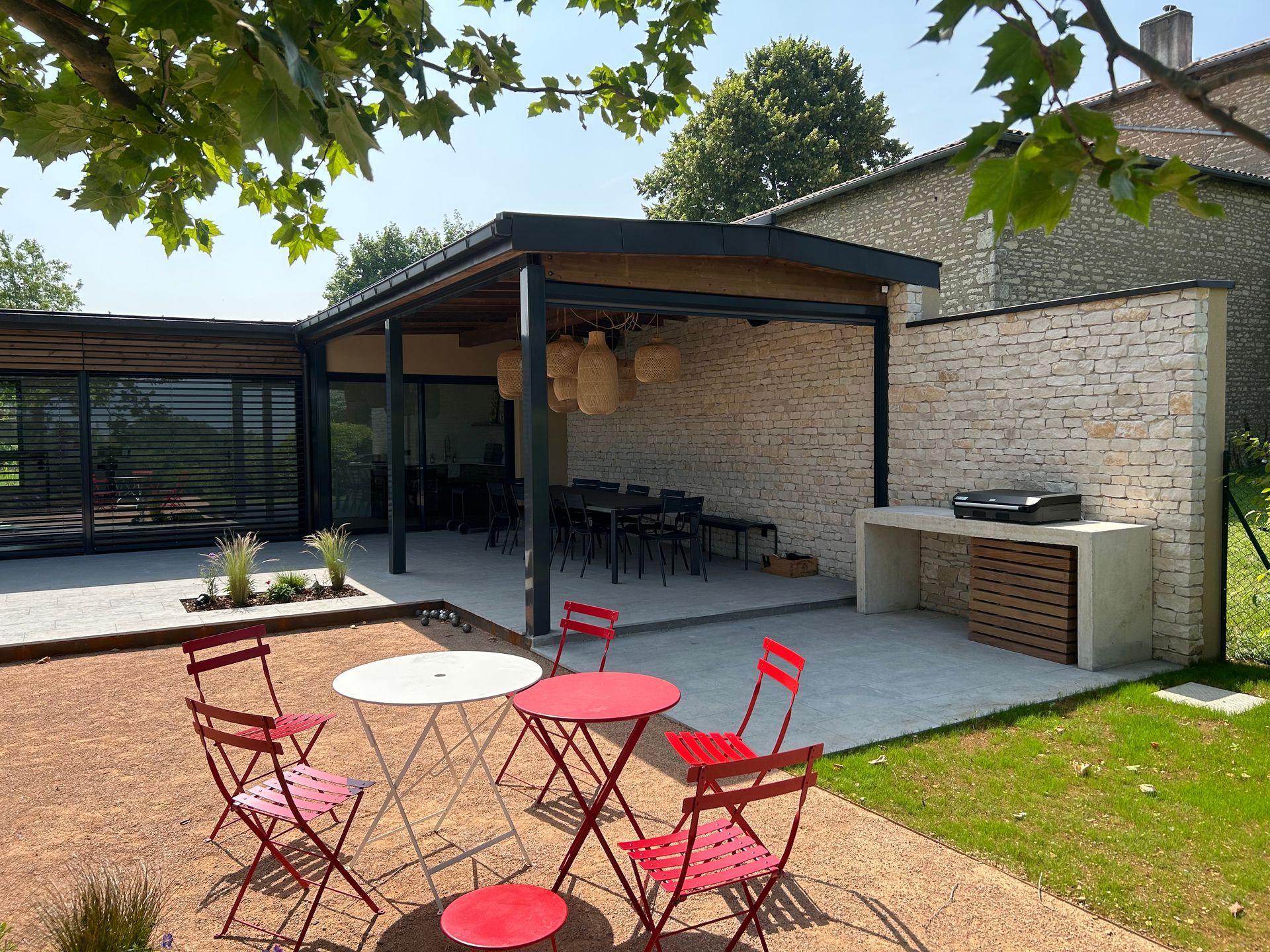 Terrasse extérieure avec chaises et table rouges, coin repas et mur en pierre avec barbecue.