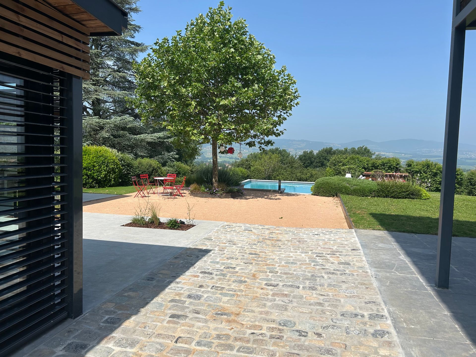 Le patio en pierre mène à une piscine et à un arbre, donnant sur un paysage ensoleillé.