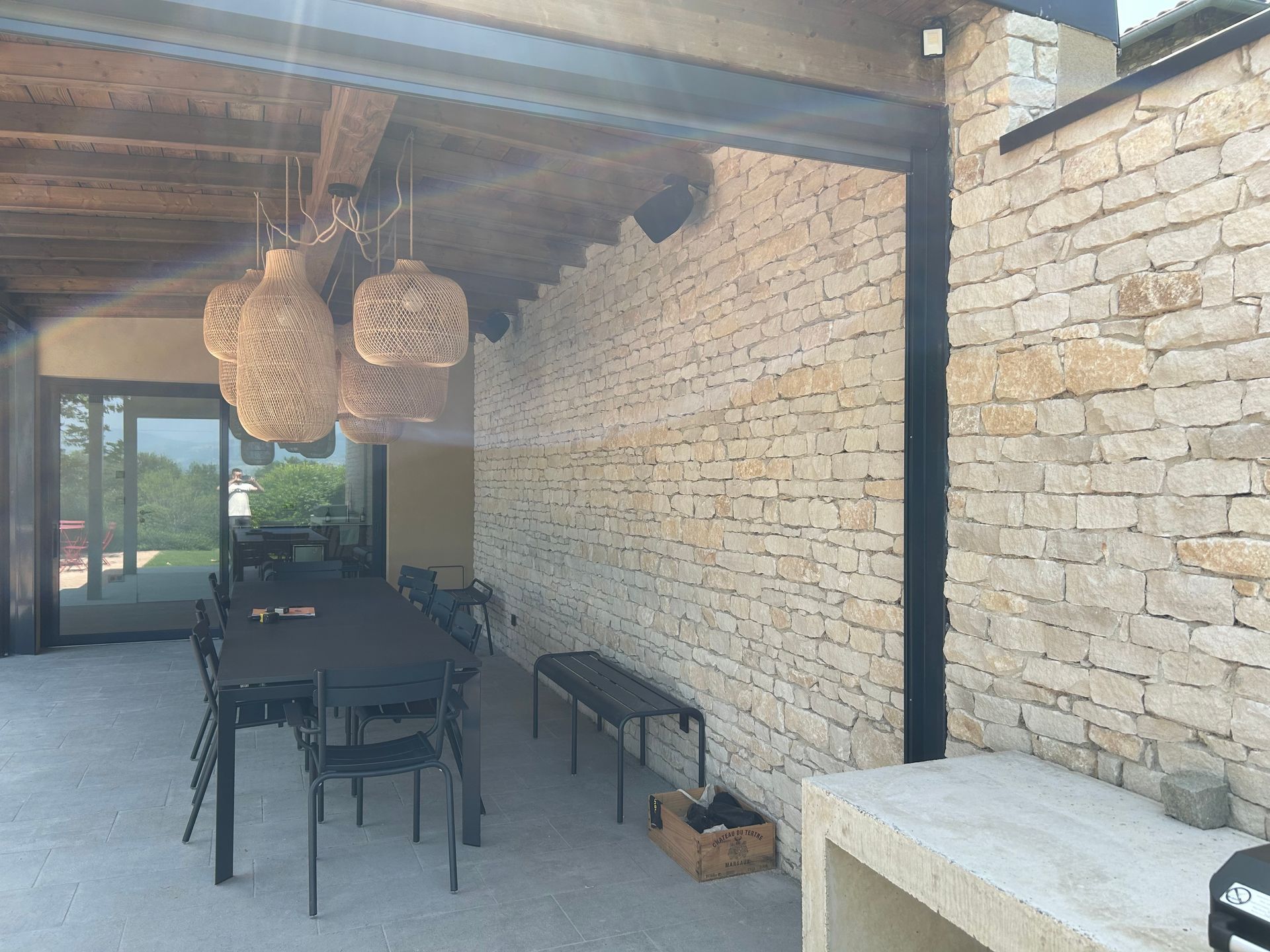 Espace repas extérieur avec mur en pierre, table et chaises noires, suspensions tissées et banc.