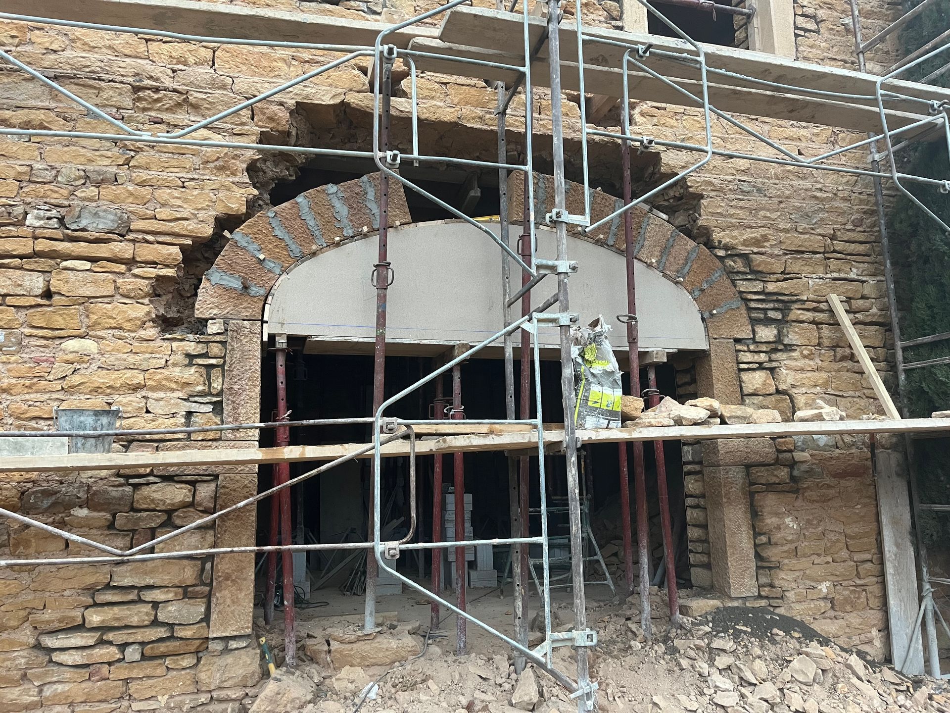 Chantier de construction avec échafaudages autour d'une ouverture en arc dans un bâtiment en pierre. On aperçoit un ouvrier en gilet.