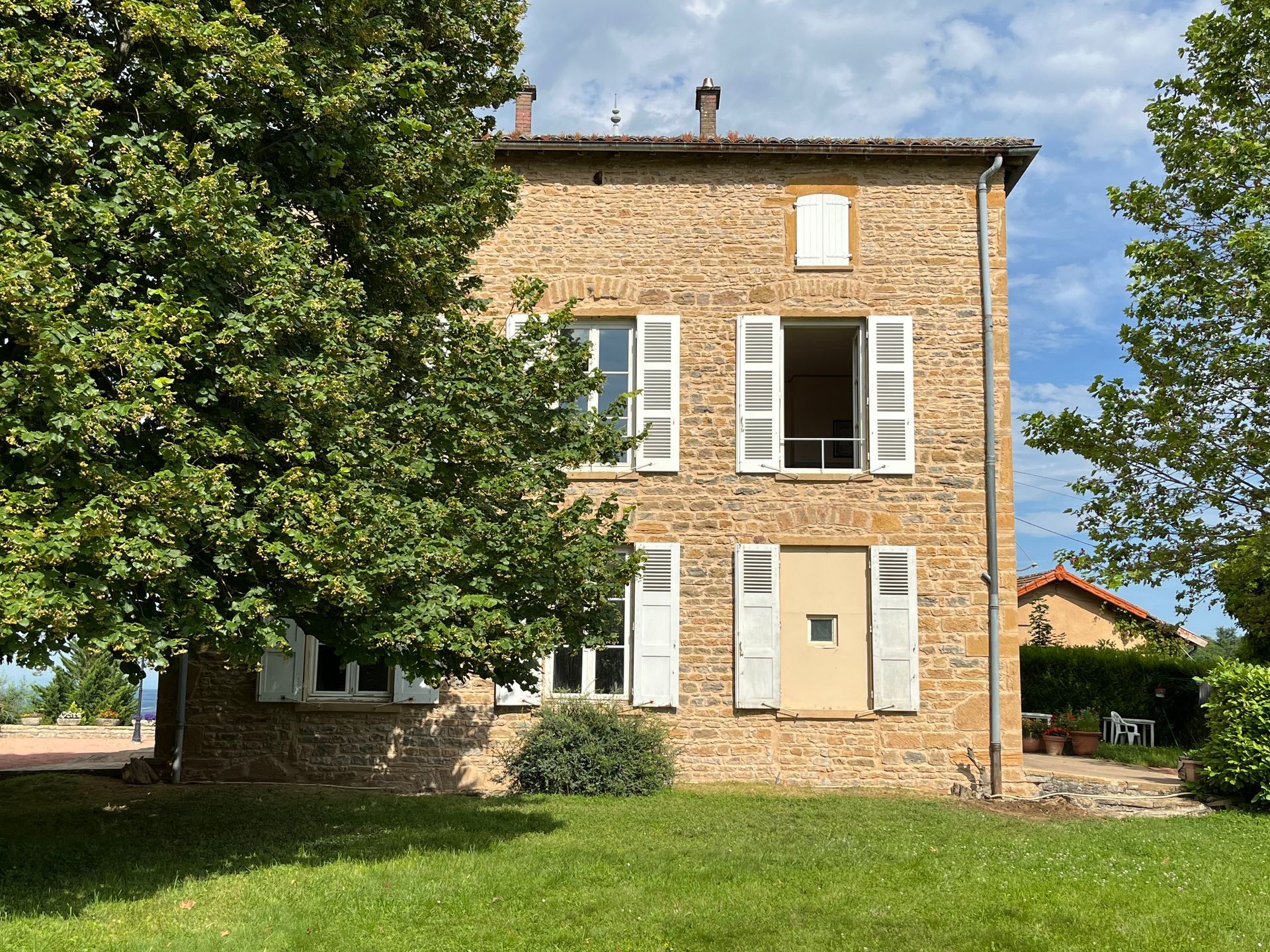 Maison en pierre avec volets blancs et fenêtre ouverte ; pelouse verte et arbres dans la cour.