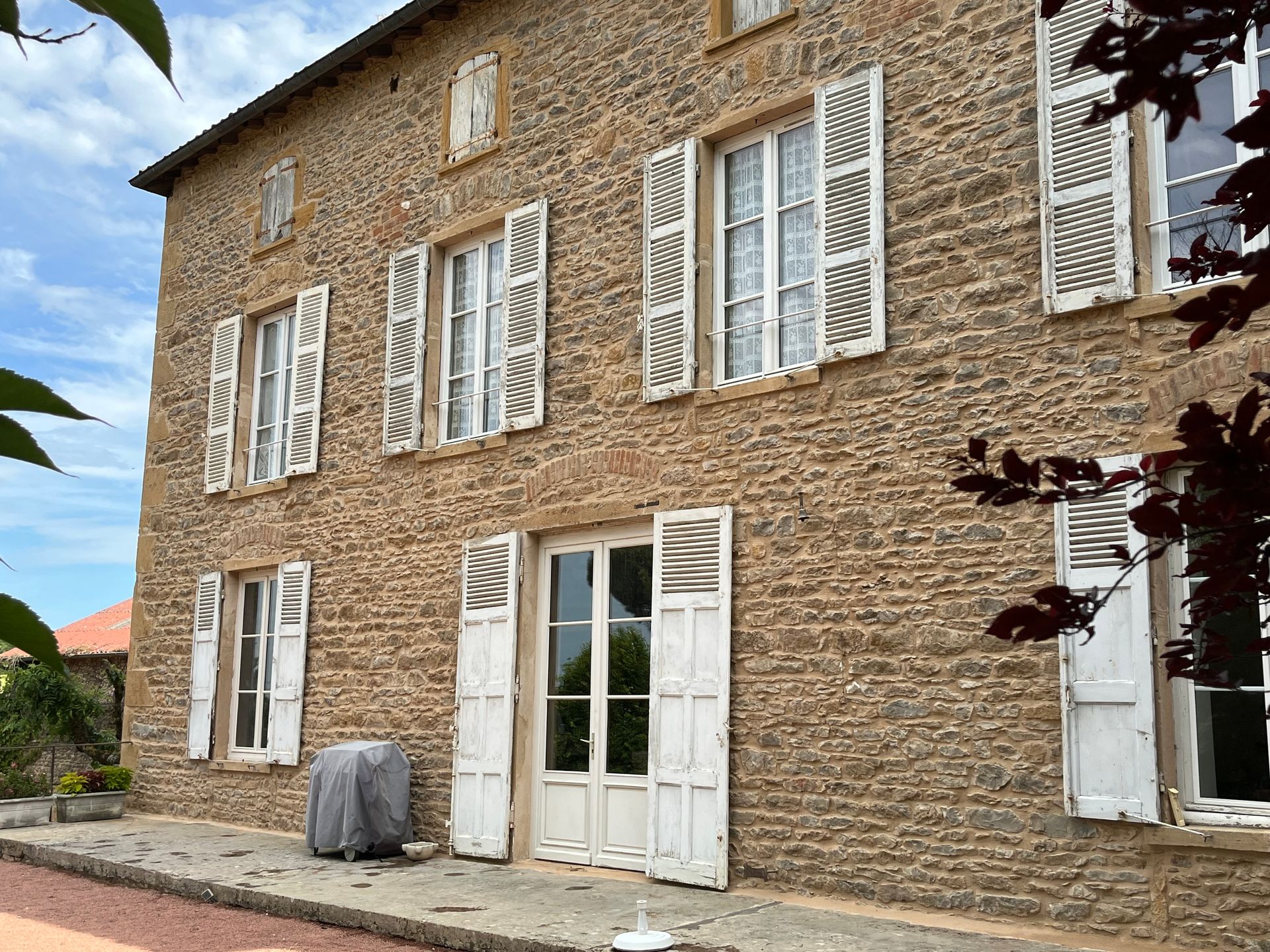 Maison en pierre avec volets et portes blancs, ciel bleu et feuillage rouge.