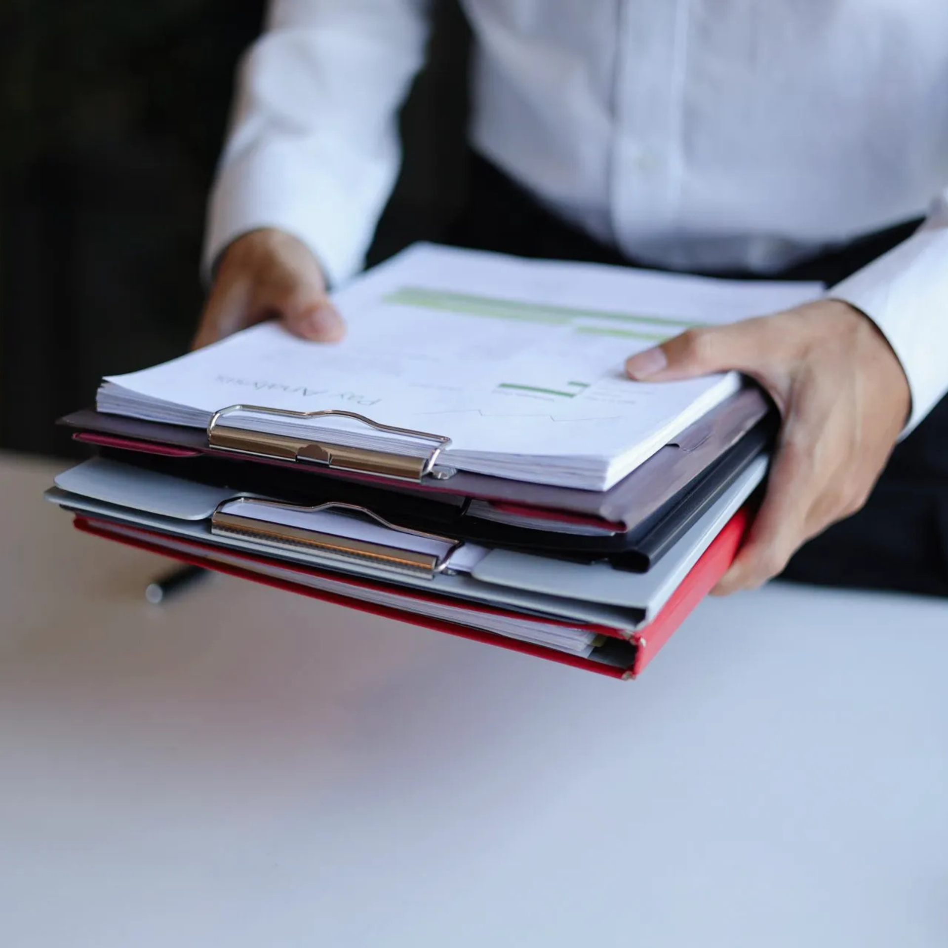 Des mains glissées dans les manches de chemises blanches tiennent une pile de porte-documents et de dossiers contre un bureau blanc.