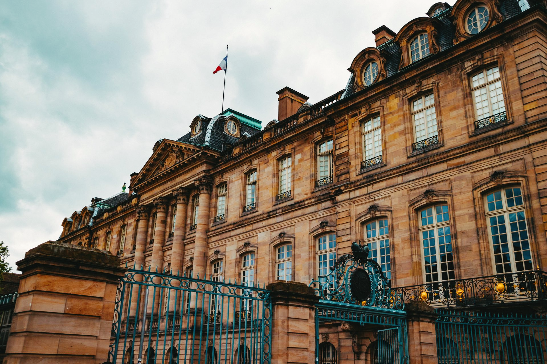 Le palais Rohan à Strasbourg, en France, orné de détails architecturaux, avec son architecture en grès rouge et sa grille en fer forgé, sous un ciel nuageux.