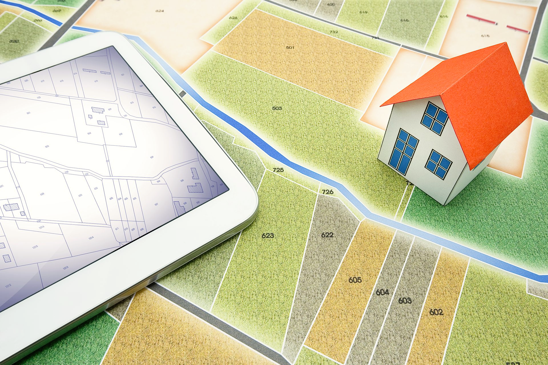 Une tablette affichant une carte est posée sur un plan de propriété imprimé, à côté d'une petite maquette 3D d'une maison.