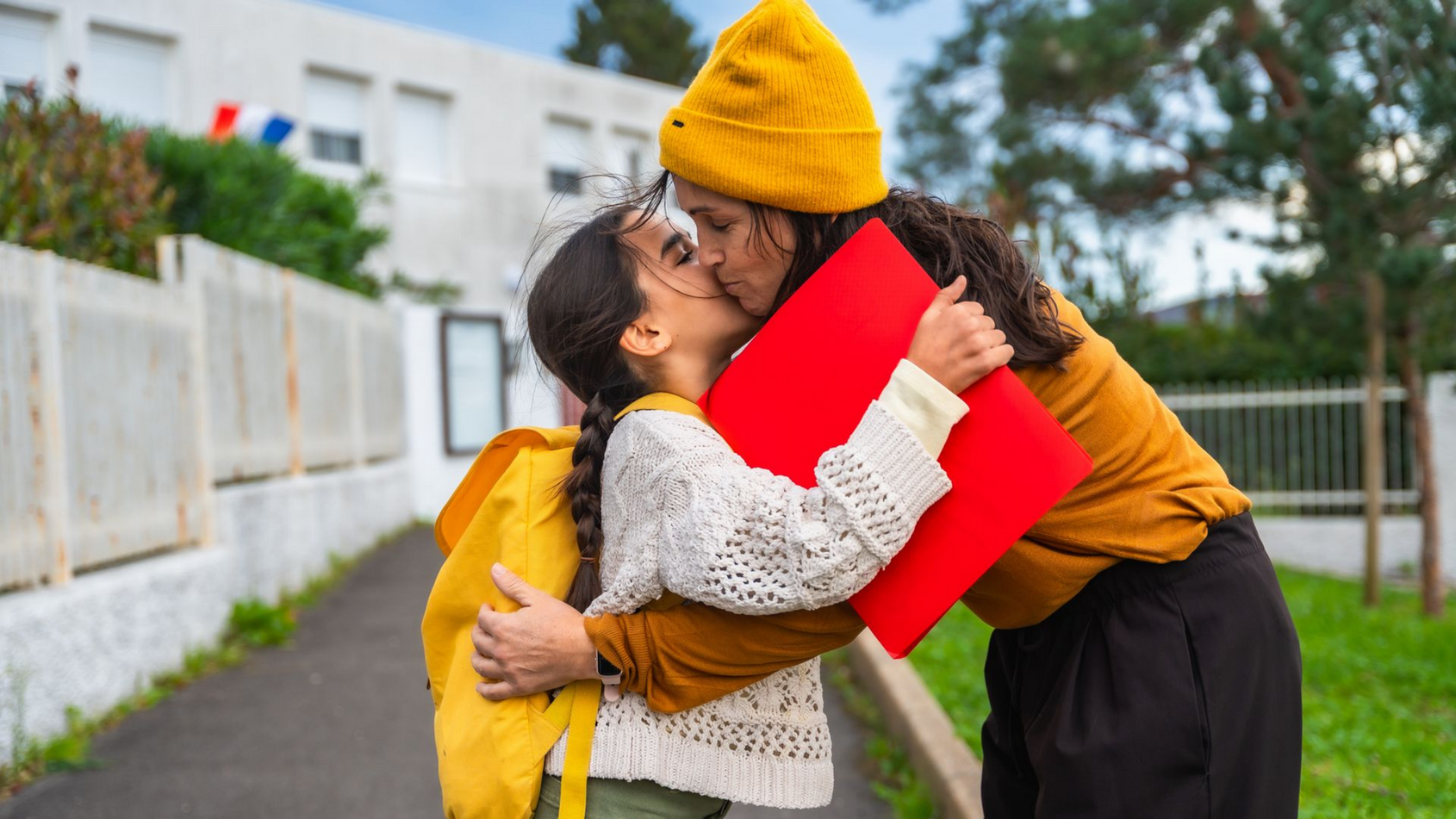 Une femme embrasse un enfant devant une école ; l'enfant porte un sac à dos et tient un classeur rouge.