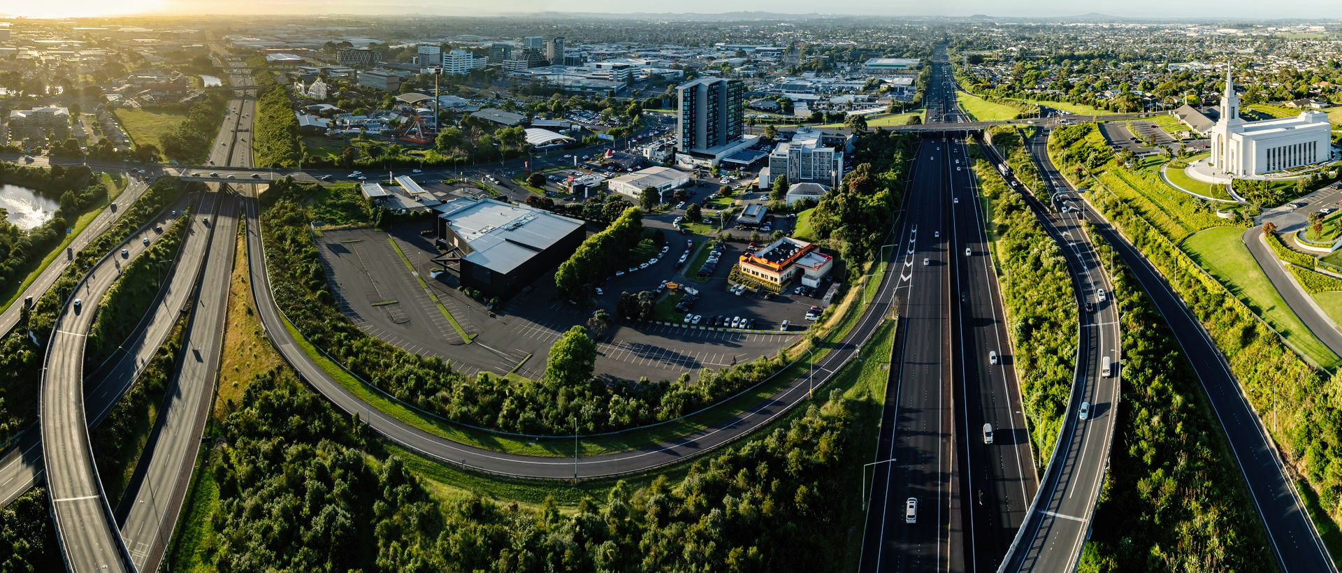 Vue aérienne d'un carrefour autoroutier à plusieurs voies entouré de verdure, de bâtiments de banlieue et d'une haute église blanche.