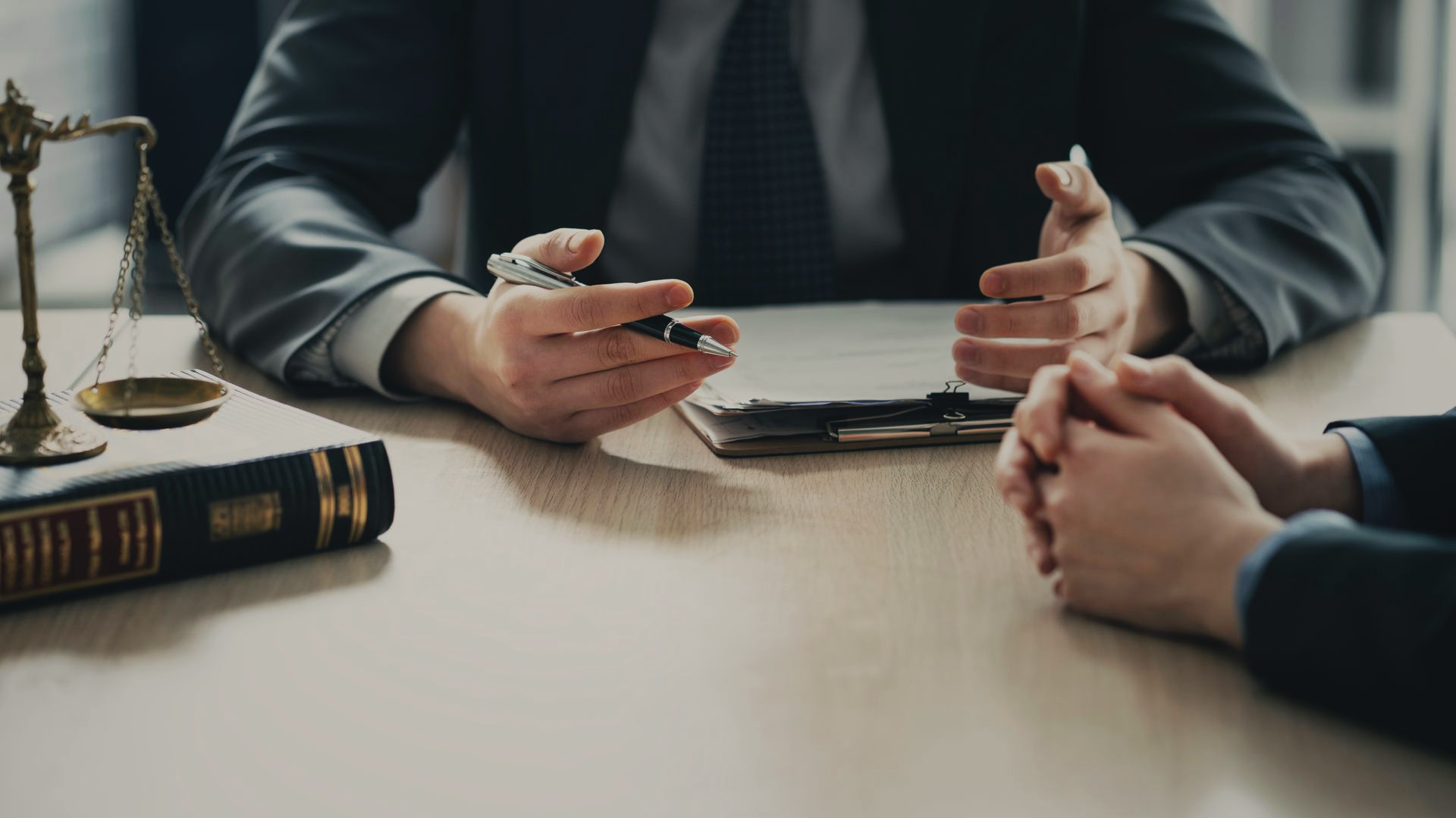 Un avocat en costume discute de documents à un bureau, à côté d'un livre et d'une balance de la justice, face à son client.