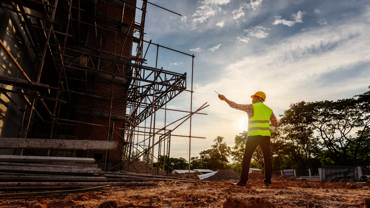 Vers page droit pénal Un ouvrier du bâtiment, vêtu d'un gilet haute visibilité et d'un casque de chantier, se tient sur un terrain vague et désigne un bâtiment en réparation.