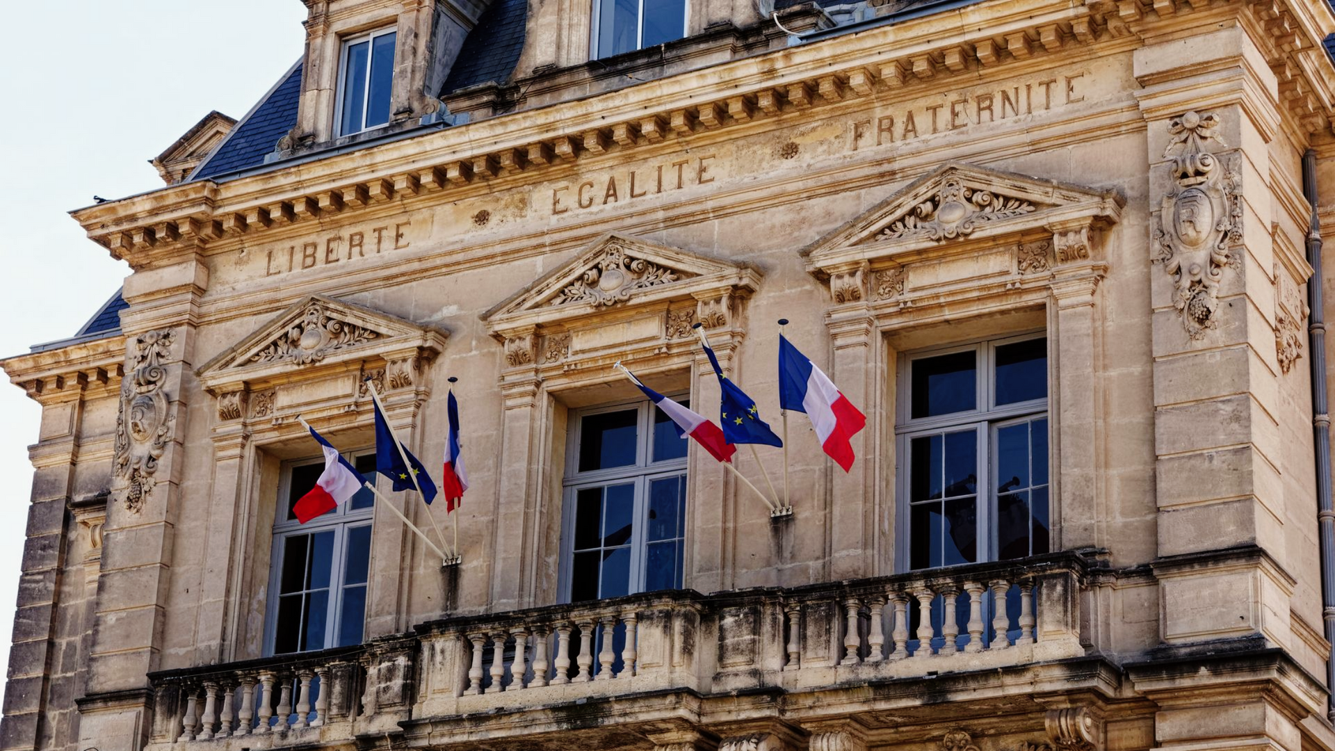 Façade d'un immeuble avec des drapeaux français et l'inscription : LIBERTÉ ÉGALITÉ FRATERNITÉ.