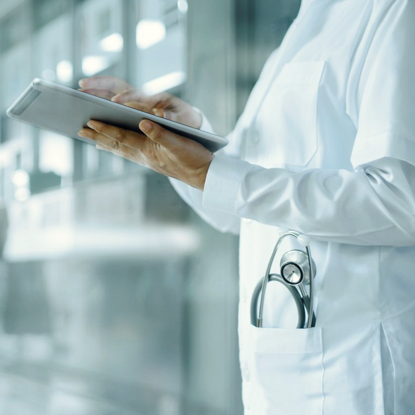 Un professionnel de santé en blouse blanche utilise une tablette dans un environnement clinique lumineux, avec un stéthoscope dans sa poche.