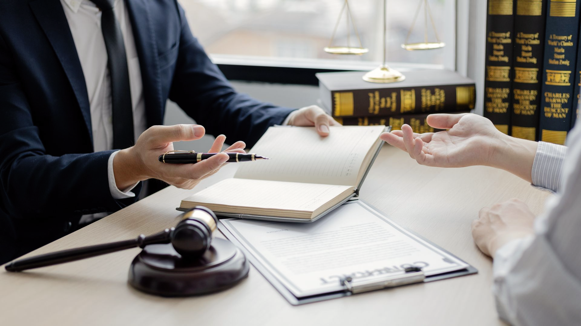 Dans un cabinet d'avocats, un avocat en costume et son client sont assis à un bureau avec un maillet, des ouvrages juridiques et un contrat.
