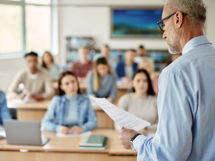 Vers page droit de l'éducation Un professeur donne un cours dans une salle de classe, en tenant des documents ; les étudiants écoutent et prennent des notes.
