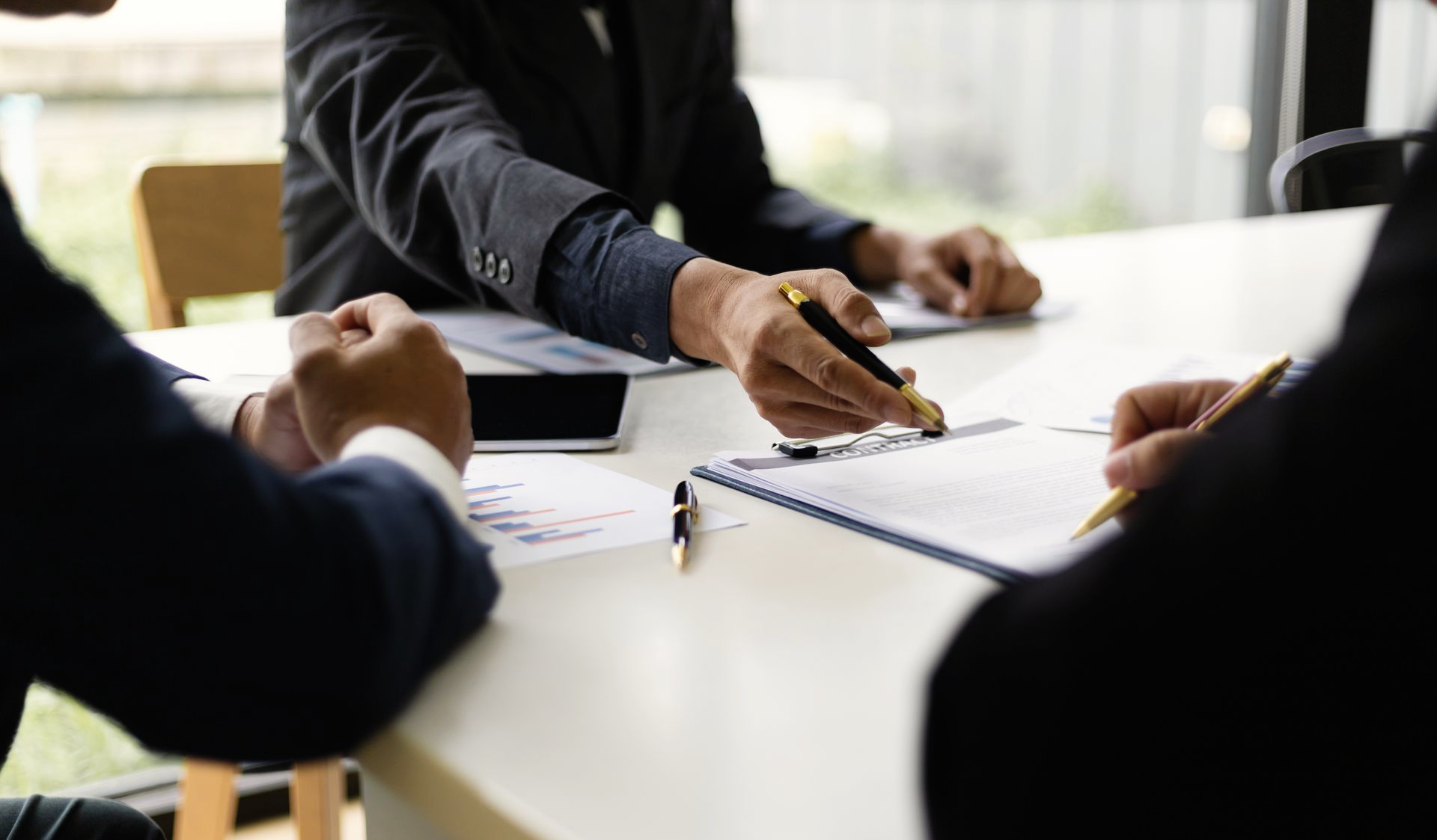 Des hommes d'affaires en costume, assis autour d'une table blanche, examinent des documents et pointent du doigt un bloc-notes.
