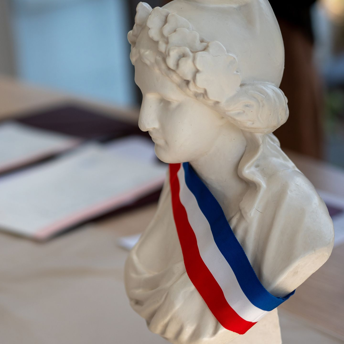 Buste d'une femme portant une écharpe tricolore, posé sur une table avec des documents.