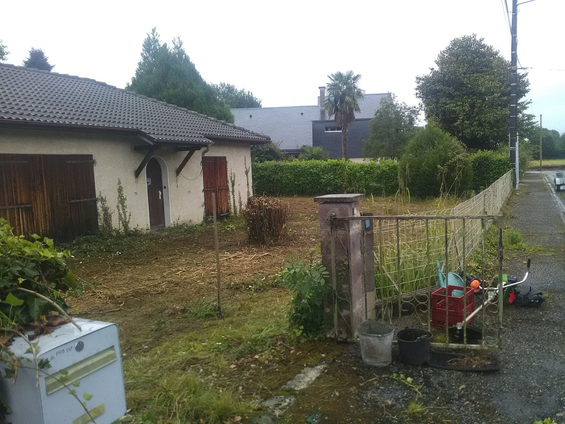 Jardin envahi par la végétation avec garage, clôture et équipement de jardinage. Ciel gris.