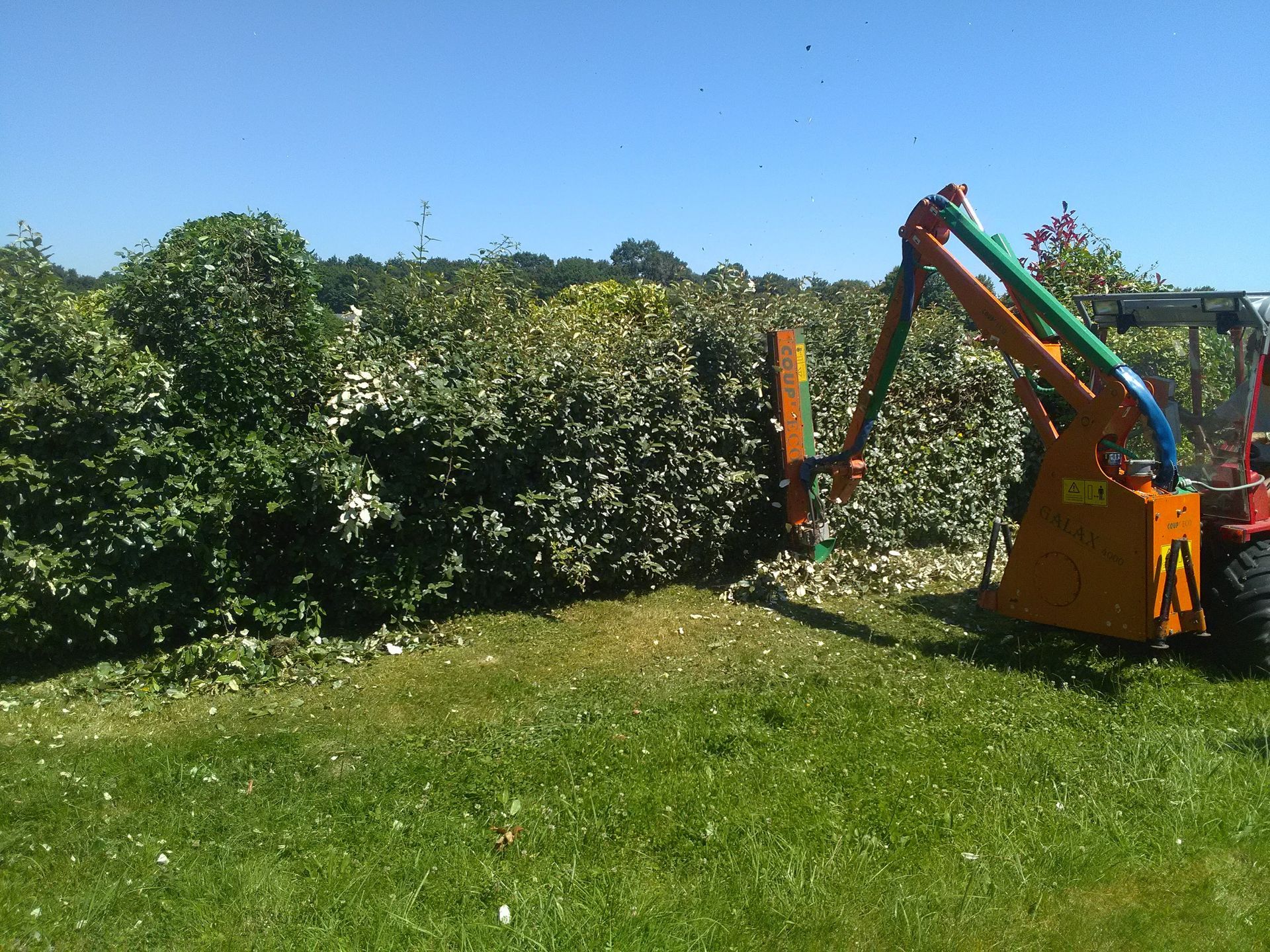 Un tracteur équipé d'un coupe-bordures monté sur bras coupe une grande haie verte dans un champ ensoleillé.