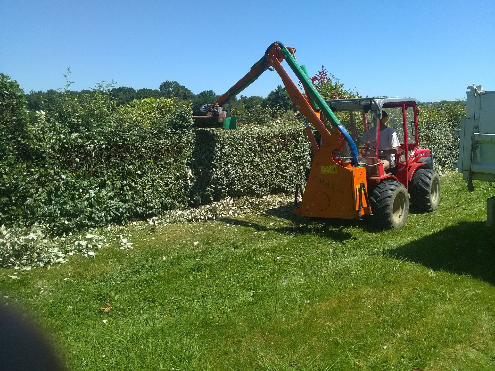 Un tracteur avec un bras étendu taille une haie, par une journée ensoleillée.