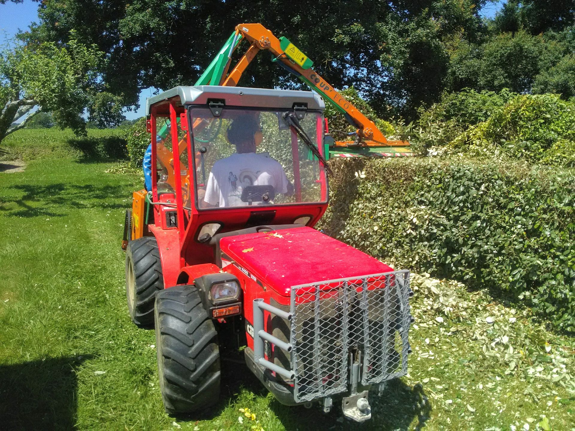 Tracteur rouge avec un taille-haie taillant des buissons par une journée ensoleillée.