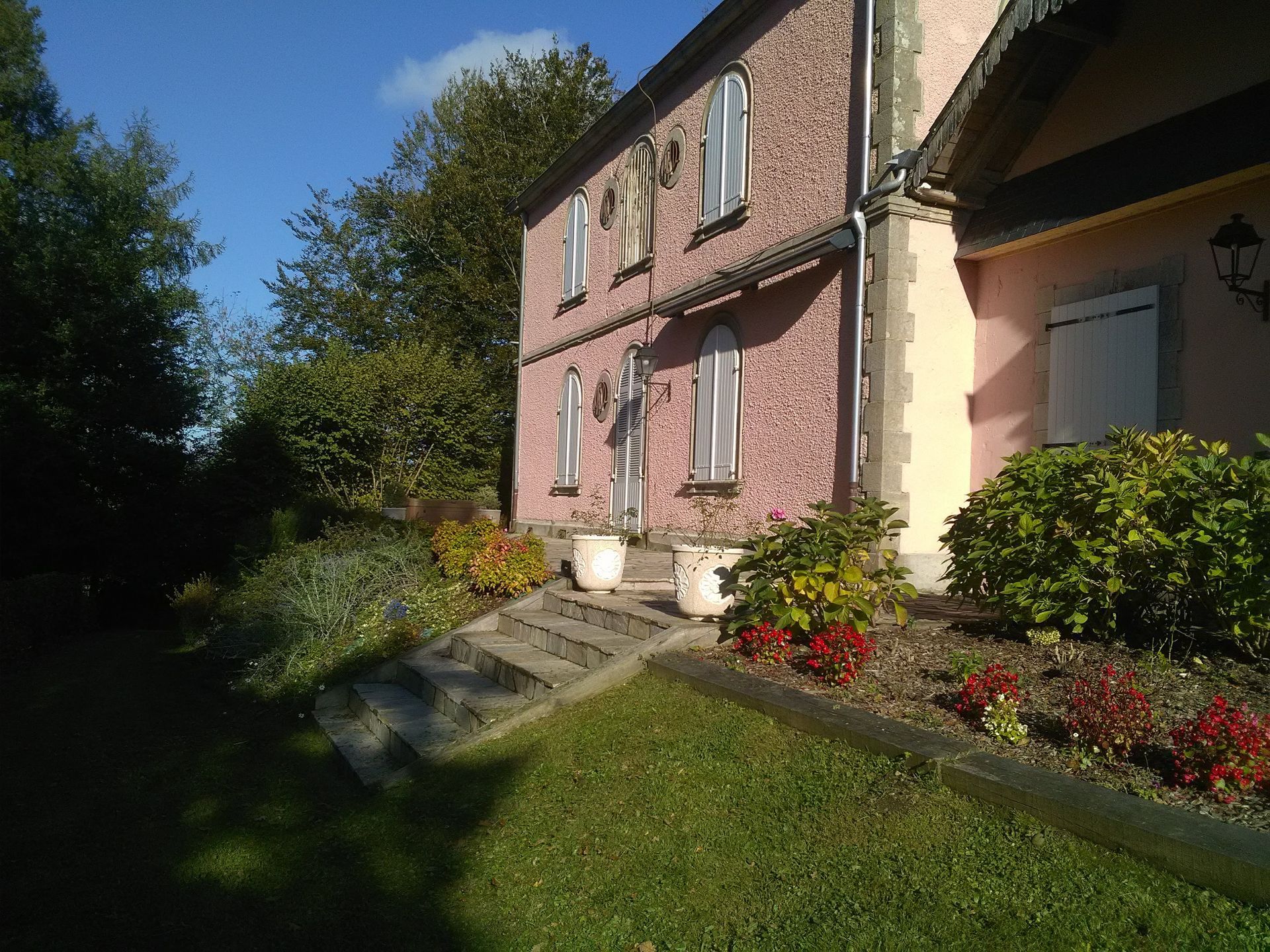 Maison rose avec des marches menant à l'entrée, entourée d'herbe verte, de buissons et de fleurs.