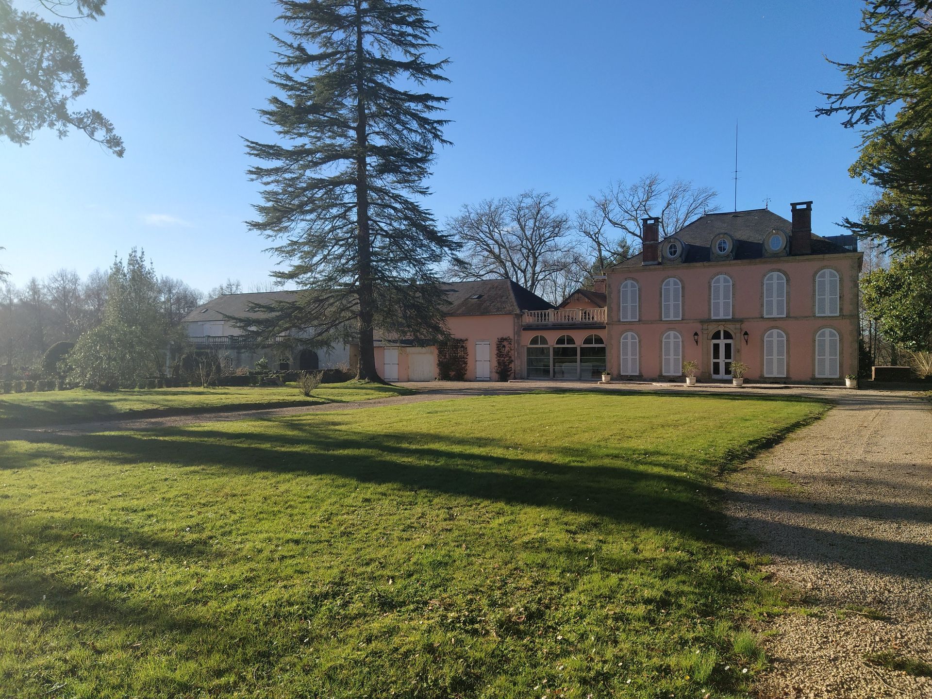 Manoir rose avec une longue allée par une journée ensoleillée, encadré par des arbres et une pelouse.