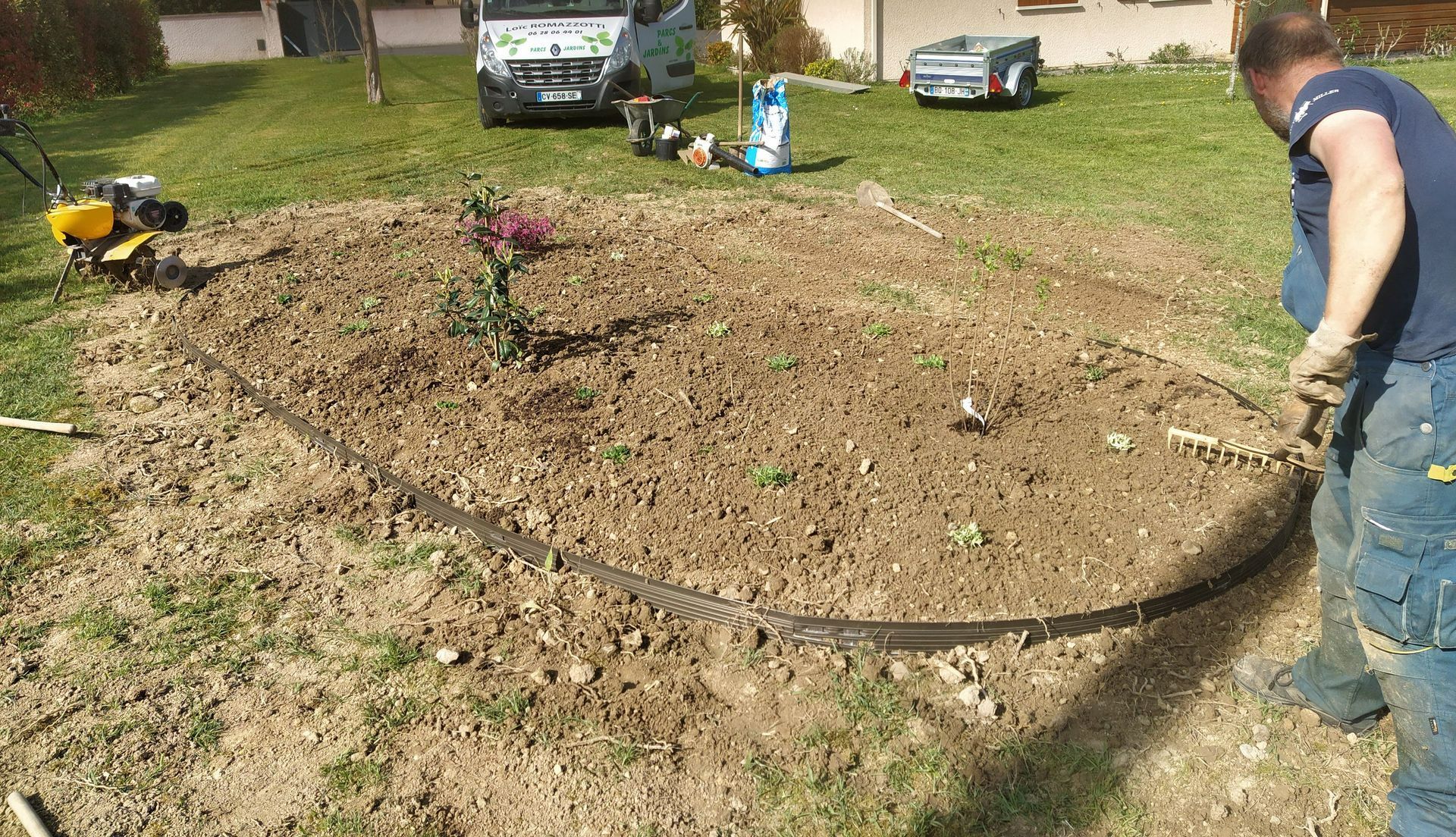 Un homme borde un lit de jardin dans une cour arrière avec de l'équipement et une camionnette visibles.