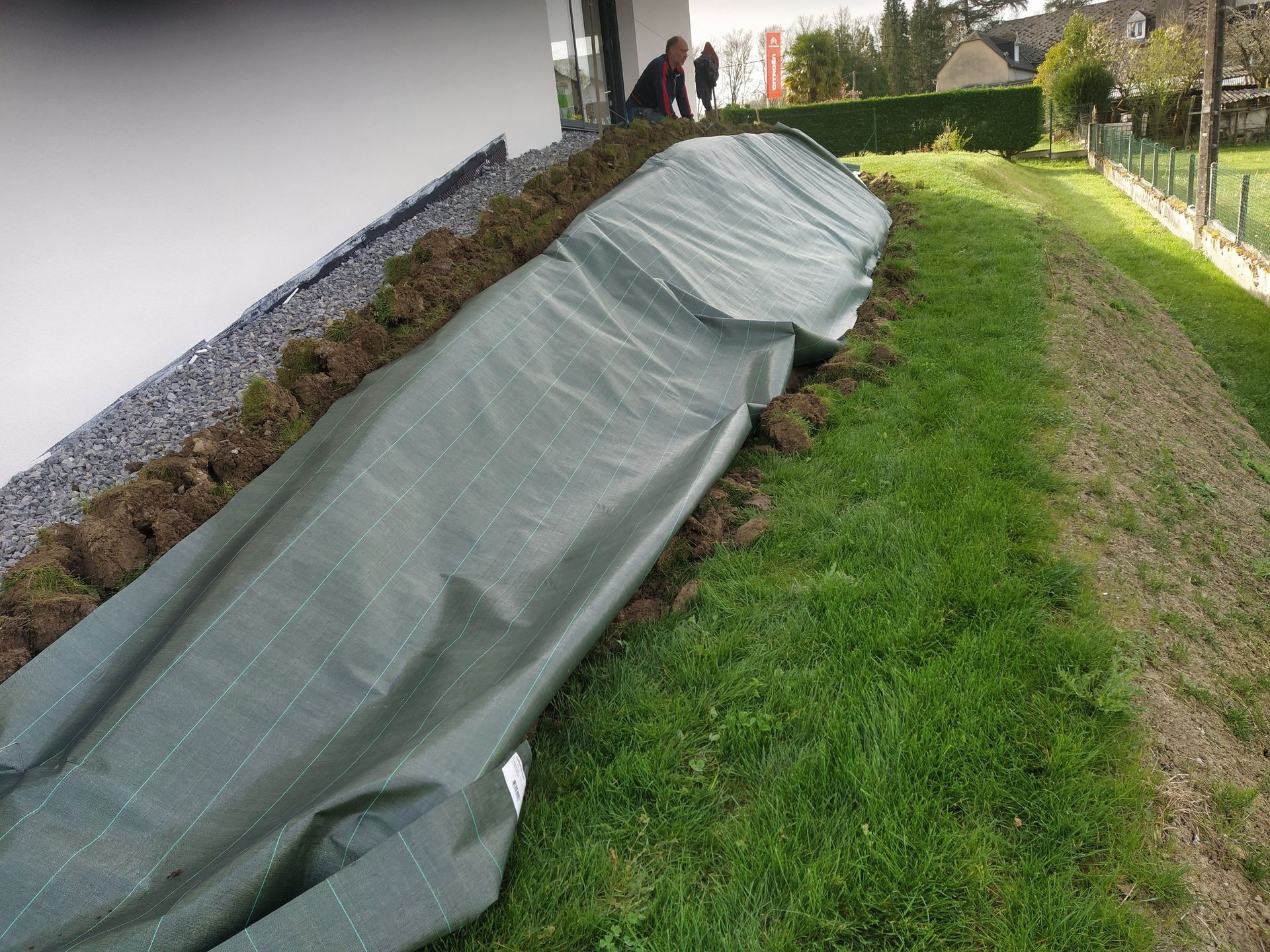 Une bâche verte recouvre une colline, près d'un bâtiment et d'une pelouse. Des personnes en arrière-plan.