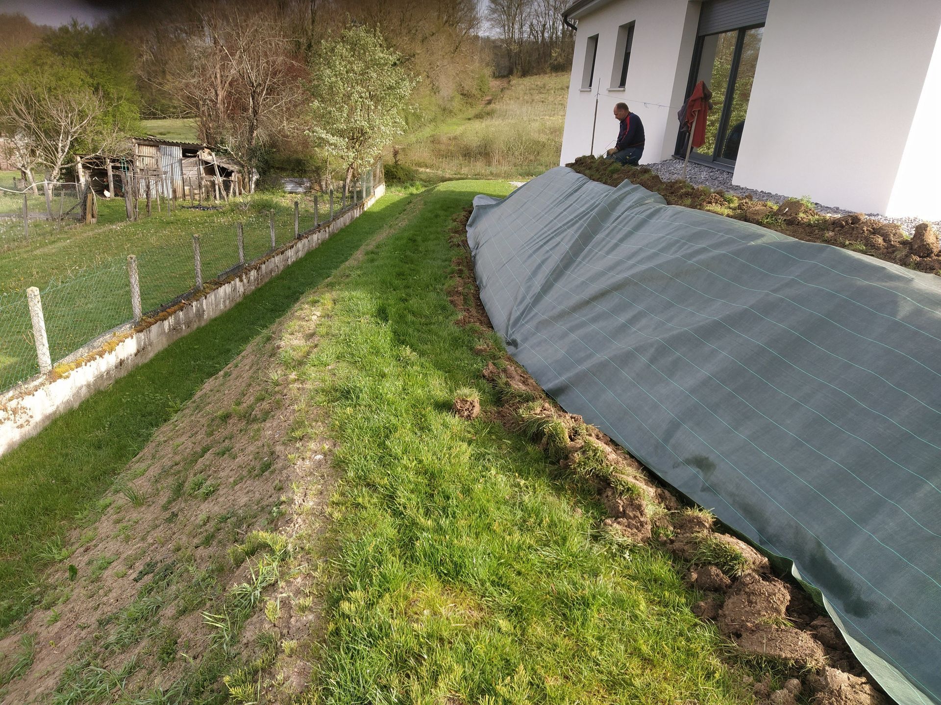 Une colline herbeuse à côté d'une maison, recouverte de tissu vert, une personne dans l'embrasure de la porte, une clôture à gauche.