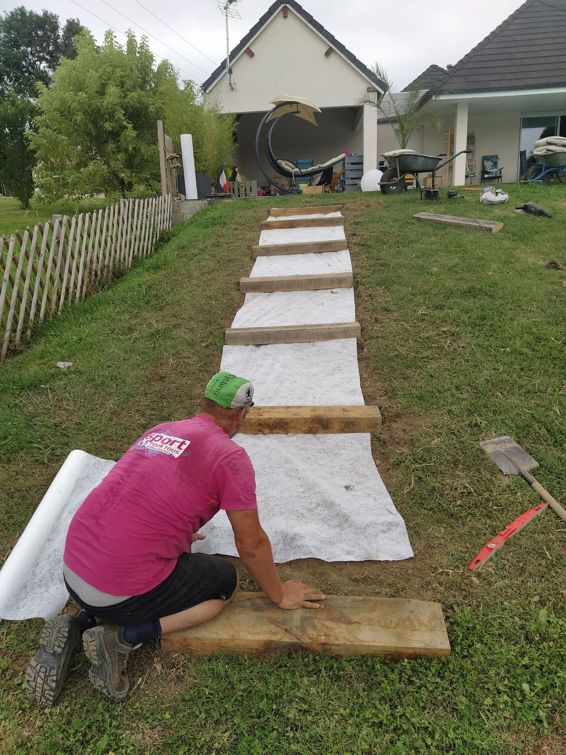 Un homme installe des marches en bois dans une cour verdoyante. Il porte une chemise rose et un chapeau vert.